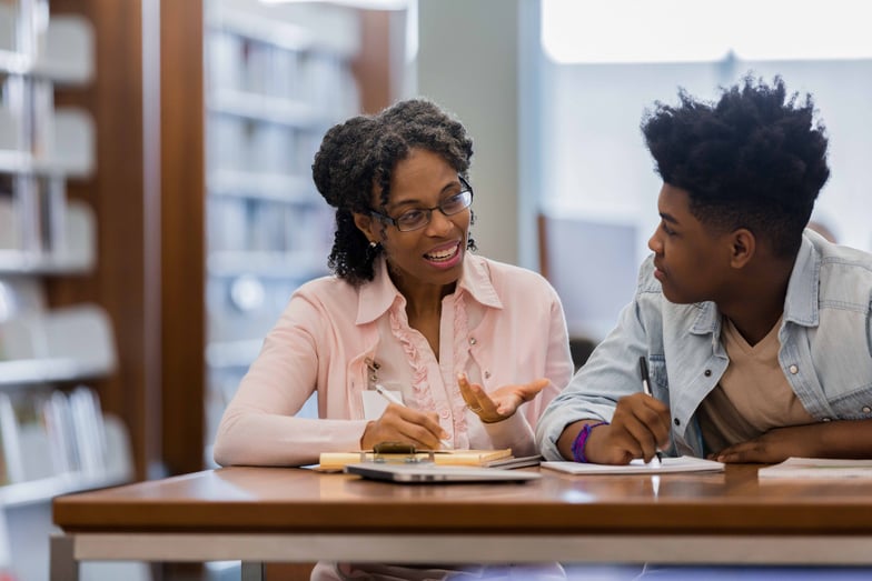 A young man and older woman are sitting at a table and talking to each other in a library.