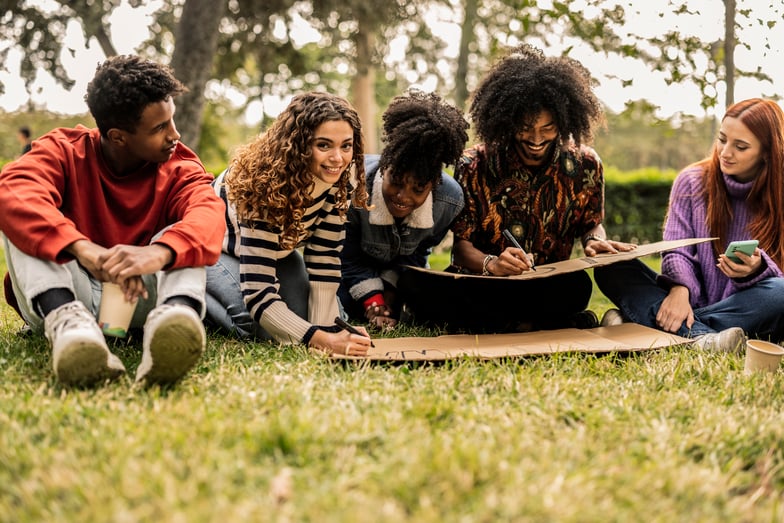 A group of young adults write on cardboard signs outdoors.
