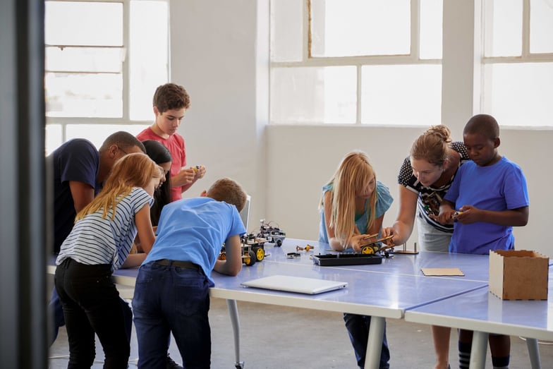 A teacher and a group of students are standing around a table working on a robotics project.
