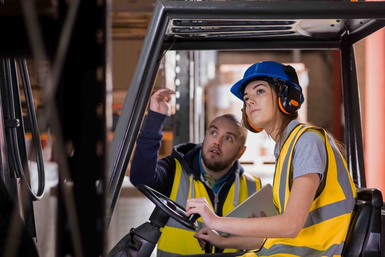 A man is teaching a young woman how to drive a forklift in a warehouse.